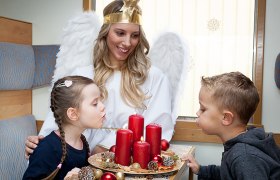 Unterwegs mit dem Christkind, © NB/citronenrot Eine Frau als Christkind verkleidet sitzt mit zwei Kindern in einem Zug. Vor ihnen steht ein Adventkranz mit roten Kerzen.