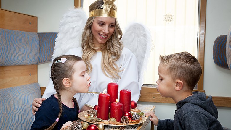 Unterwegs mit dem Christkind, © NB/citronenrot Eine Frau als Christkind verkleidet sitzt mit zwei Kindern in einem Zug. Vor ihnen steht ein Adventkranz mit roten Kerzen.