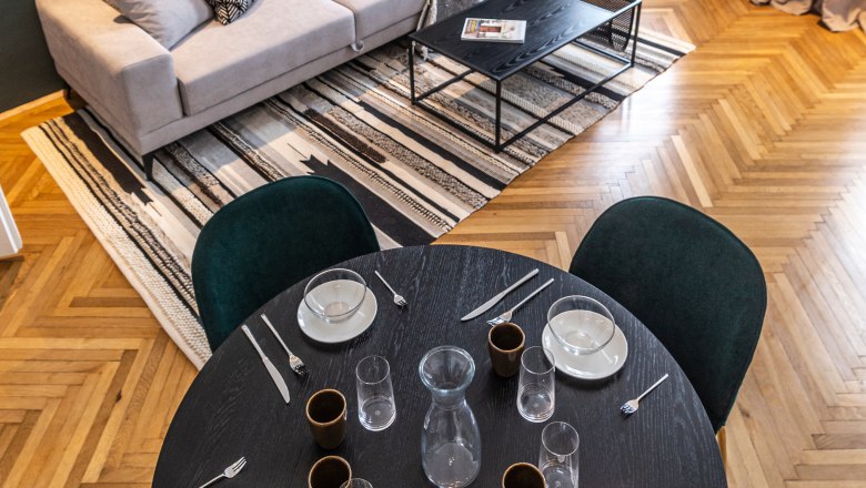 Apartment, © Astrid Bartl Modernes Wohnzimmer mit Sofa, Couchtisch, Teppich und Esstisch mit Gedeck.