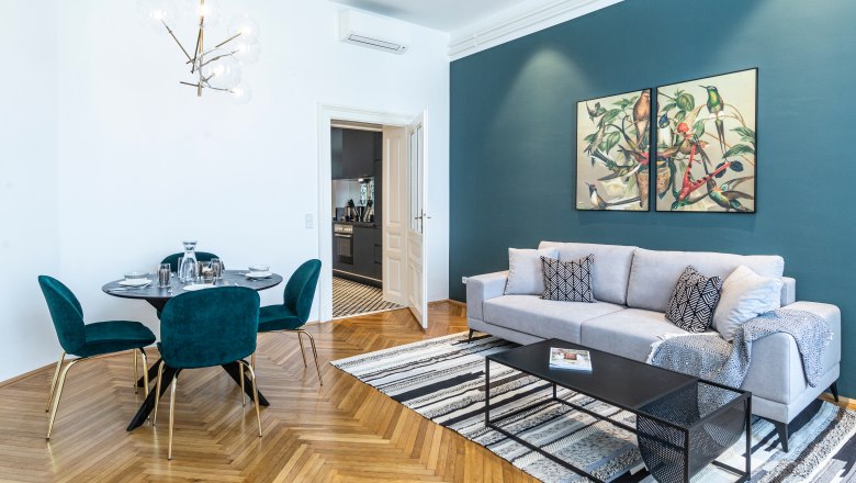 Apartment, © Astrid Bartl Modernes Wohnzimmer mit grauem Sofa, Esstisch und Kunst an der Wand.