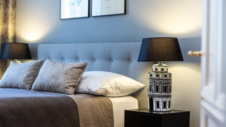 Doppelzimmer, © Astrid Bartl Elegantes Doppelzimmer mit grauem Kopfteil, zwei Kissen, Nachttischlampe und Wandbildern.