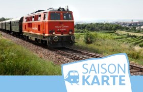 Saisonkarte Reblaus Express, © NÖVOG/Weinfranz Ein roter Zug fährt durch eine ländliche Landschaft mit Feldern und einem Dorf im Hintergrund. Unten rechts ist das Logo 'Saisonkarte' zu sehen.