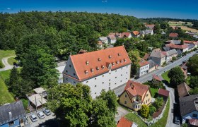 Hotel Schüttkasten, © Franz Gleiss Luftaufnahme eines großen Gebäudes mit rotem Dach, umgeben von Bäumen und kleineren Häusern in einer ländlichen Gegend.