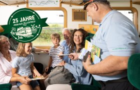 Eine Familie sitzt in einem Zugabteil des Reblaus Express und wird von einem Schaffner bedient.