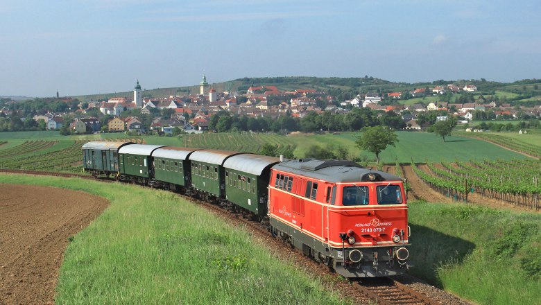 Zug f&auml;hrt vorbei an Weinreben. Im Hintergrund sieht man eine Ortschaft.