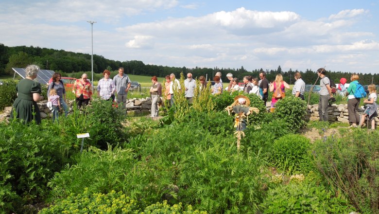 Menschen bei einem Fest in einem Bauerngarten mit Vogelscheuche und Pflanzen.