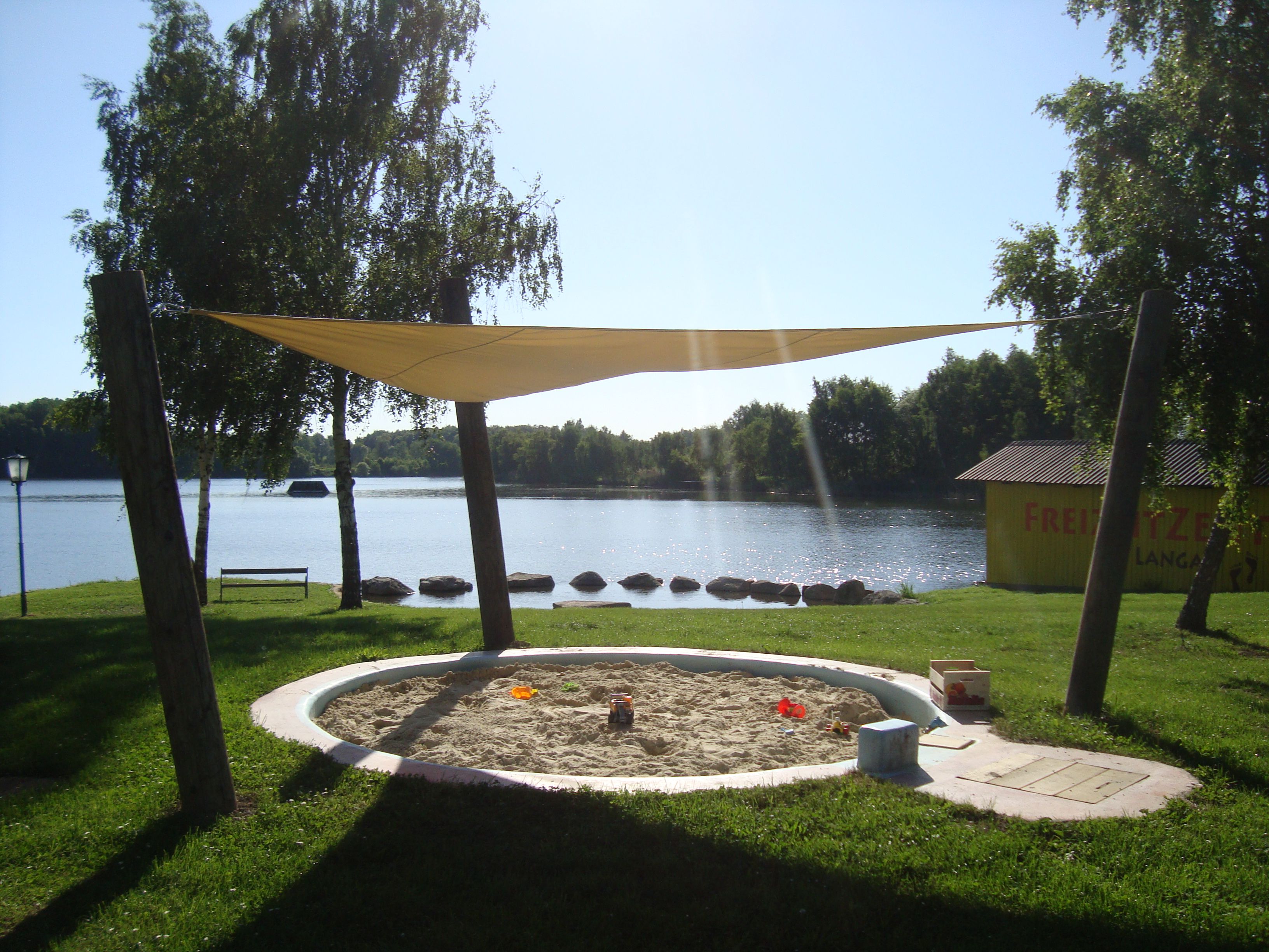 Ein Sandkasten mit Sonnensegel am Ufer eines Sees, umgeben von Bäumen und einer Wiese. Im Hintergrund ein gelbes Gebäude mit der Aufschrift 'Freizeitzentrum Langau'.