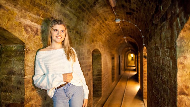 Eine Frau in einem weißen Pullover steht in einem beleuchteten, gemauerten Keller.