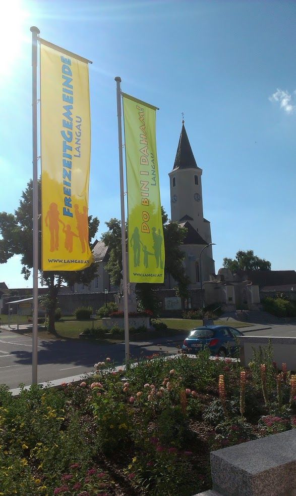Zwei bunte Fahnen mit der Aufschrift 'Freizeitgemeinde Langau' und 'Do bin i daham' vor einer Kirche.