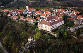 Luftaufnahme eines Dorfes mit roten Dächern, Kirche mit Turm und einem großen Schloss auf einem bewaldeten Hügel
