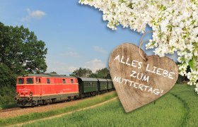 Roter Zug f&auml;hrt durch gr&uuml;ne Landschaft, daneben ein Holzherz mit der Aufschrift 'Alles Liebe zum Muttertag' und wei&szlig;en Bl&uuml;ten im Hintergrund.
