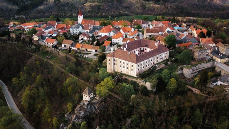 Luftaufnahme eines Dorfes mit roten Dächern, Kirche mit Turm und einem großen Schloss auf einem bewaldeten Hügel