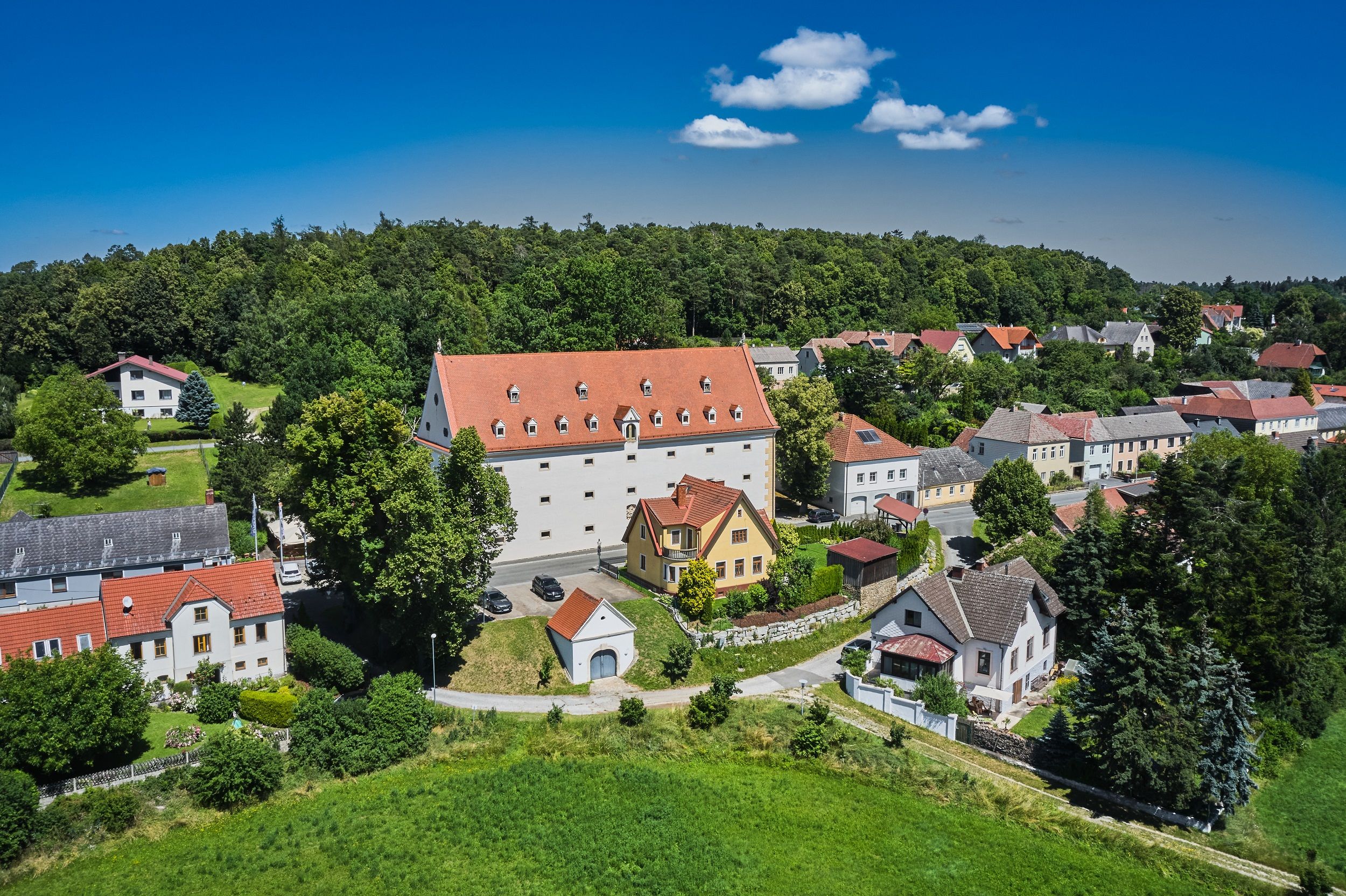 Luftaufnahme eines Dorfes mit einem großen weißen Gebäude mit rotem Dach, umgeben von kleineren Häusern und viel Grün.