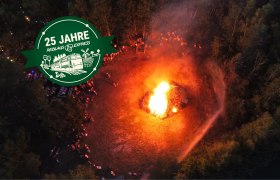 Luftaufnahme eines gro&szlig;en Sonnwendfeuer in einem Waldgebiet mit Menschen drumherum.