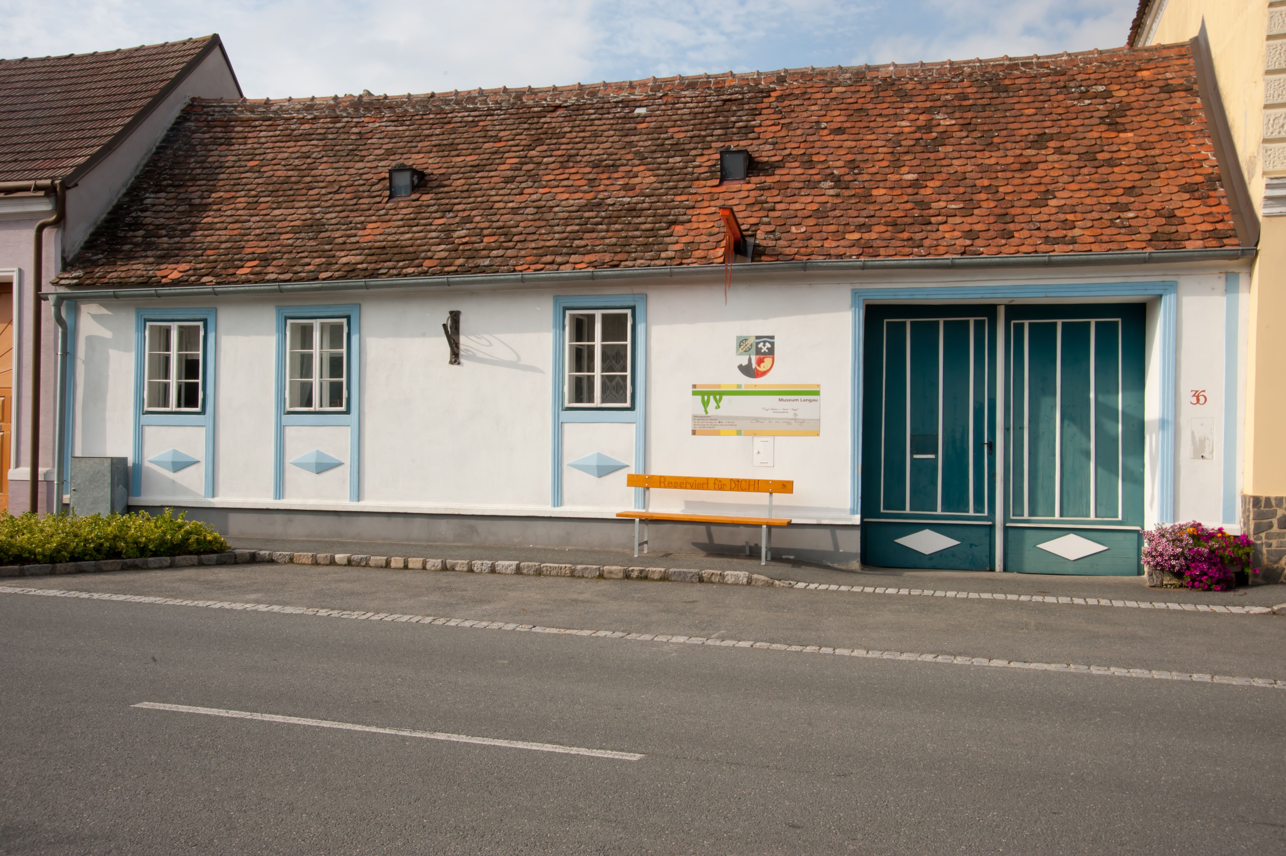 Ein kleines, traditionelles Gebäude mit roten Ziegeln und blauen Fensterrahmen. Vor dem Gebäude steht eine gelbe Bank mit der Aufschrift 'Reserviert für DICH!'.