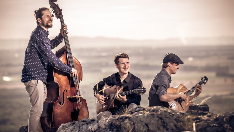 Drei Musiker spielen Kontrabass und Gitarren im Freien vor weiter Landschaft bei Sonnenuntergang.