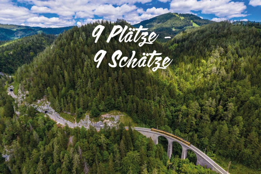Bahn fährt über ein Viadukt durch bewaldete Berge unter blauem Himmel mit Wolken, Text '9 Plätze - 9 Schätze' in der Bildmitte
