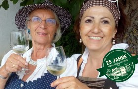 Zwei Frauen in traditioneller Kleidung sto&szlig;en mit Weingl&auml;sern an. Ein Logo mit der Aufschrift '25 Jahre Reblausexpress' ist eingeblendet.