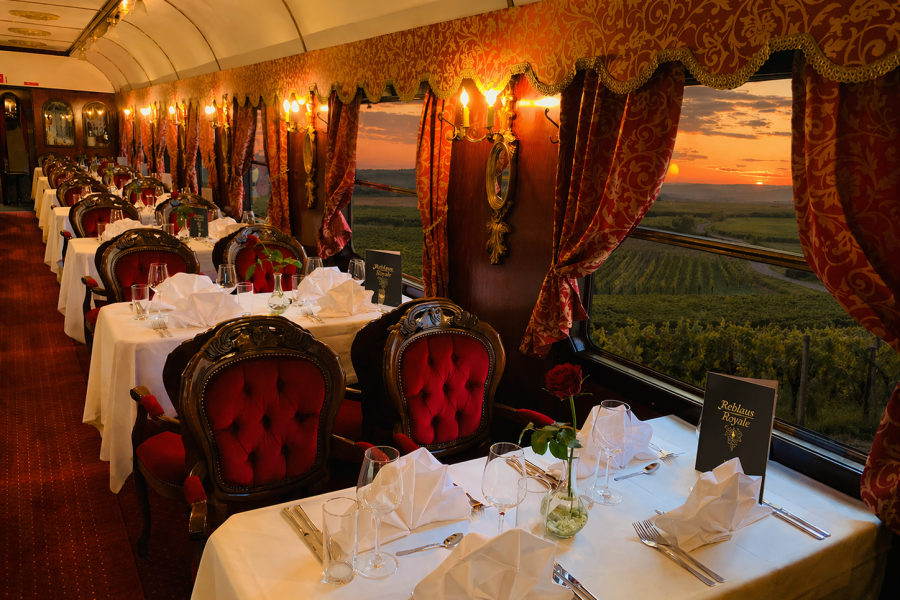 Eleganter Speisewagen eines Zuges mit rot-goldener Einrichtung, gedeckten Tischen und gepolsterten St&uuml;hlen; durch die gro&szlig;en Fenster ist eine Landschaft mit Weinbergen bei Sonnenuntergang zu sehen.