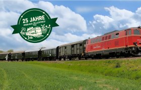 Roter Zug mit mehreren Waggons f&auml;hrt auf Gleisen durch eine gr&uuml;ne Landschaft unter blauem Himmel, daneben gr&uuml;nes Jubil&auml;umslogo